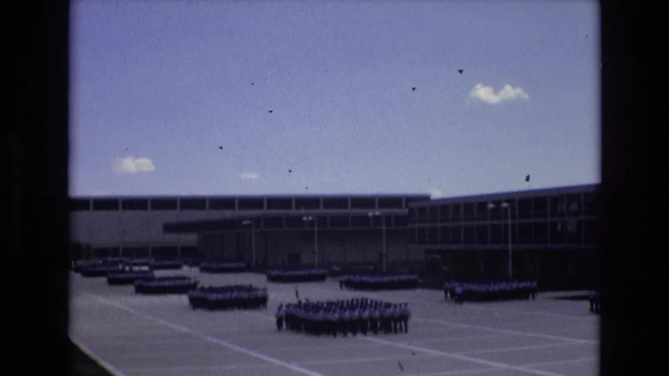 The home-movie view shows a central group of uniformed personnel clustered in rows on the plaza, with additional ranks and the institutional facade visible behind them.