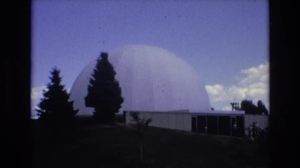 A white geodesic dome appears behind dark evergreen trees and a low building entrance, matching the supplied Colorado Springs 1973 campus architecture context.