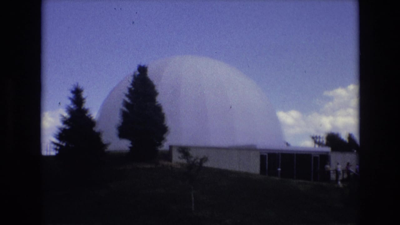 1973 Colorado Springs 8mm frame showing a large white domed building above a low entrance structure, with dark evergreen trees and bright clouds in the background.