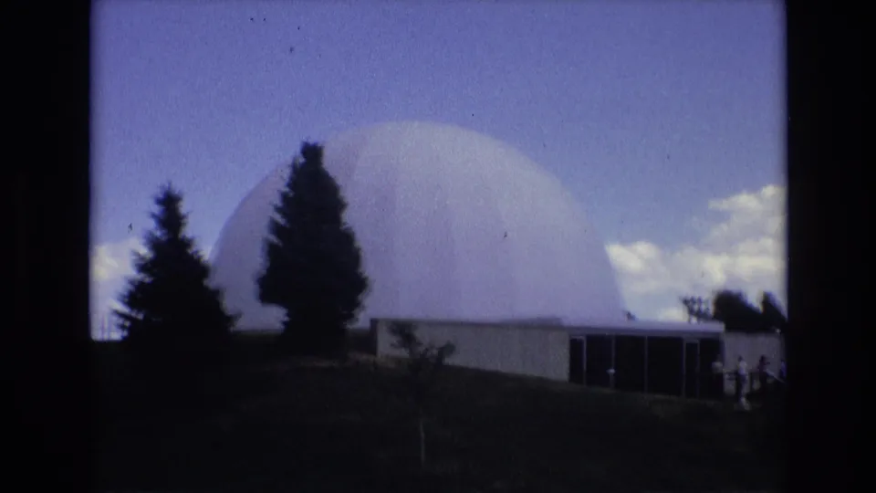 1973 Colorado Springs 8mm frame showing a large white domed building above a low entrance structure, with dark evergreen trees and bright clouds in the background.