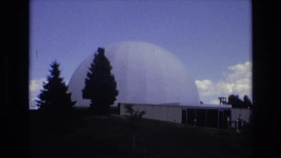 1973 Colorado Springs home-movie frame of a white domed structure partly framed by evergreen trees, with a low modern facade and small visitors near the entrance area.