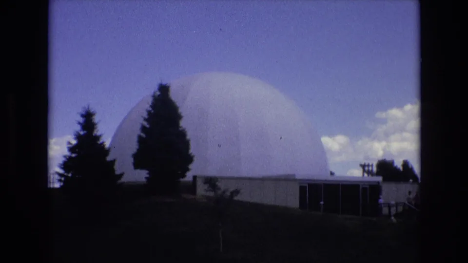 1973 Colorado Springs 8mm frame showing the domed building from a slightly lower angle, with evergreen silhouettes, a low entrance structure, and clouds along the right side of the sky.