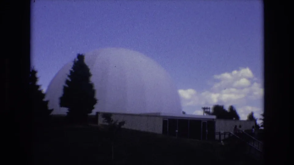 1973 Colorado Springs home-movie frame of a large white dome over a low building, with evergreen trees, a dark sloped foreground, bright clouds, and a railing or walkway at the right edge.