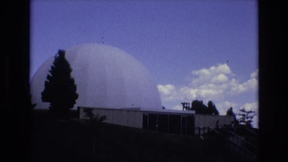 1973 Colorado Springs 8mm frame showing a large white dome rising behind a low building, with silhouetted trees and clouds in the sky.