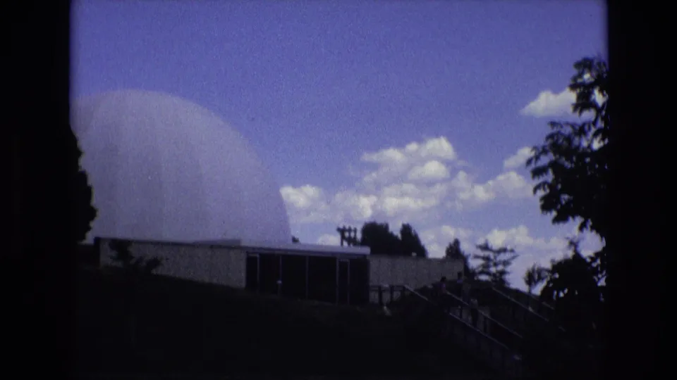 1973 Colorado Springs 8mm frame showing the dome partly cropped at left, the low building below, exterior stairs, trees, and bright clouds.