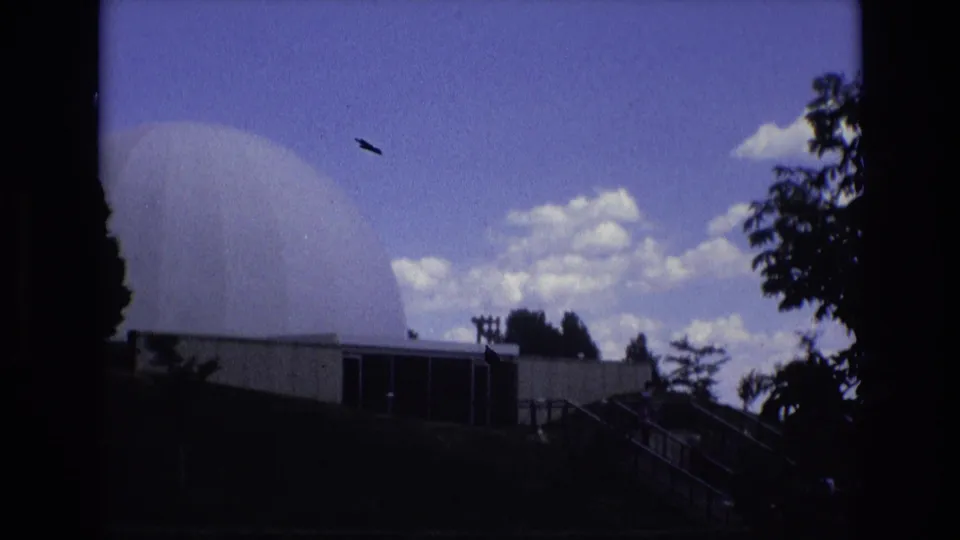 1973 Colorado Springs home-movie frame with the white dome, building, stair railings, silhouetted trees, clouds, and a small dark speck or possible aircraft in the sky.