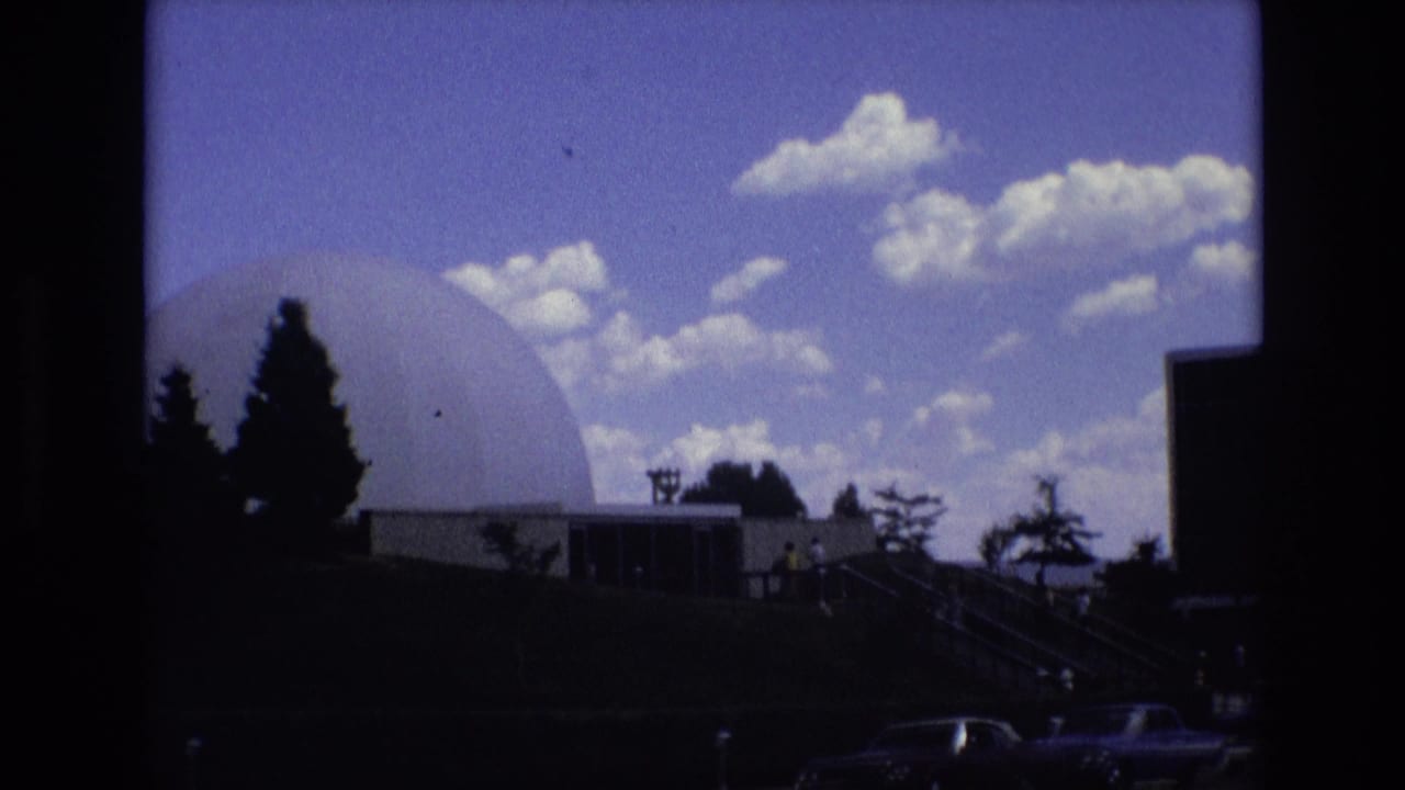 1973 Colorado Springs home-movie frame showing a large white geodesic dome near dark building sections, silhouetted evergreens, stairs, cars, and a bright clouded sky.