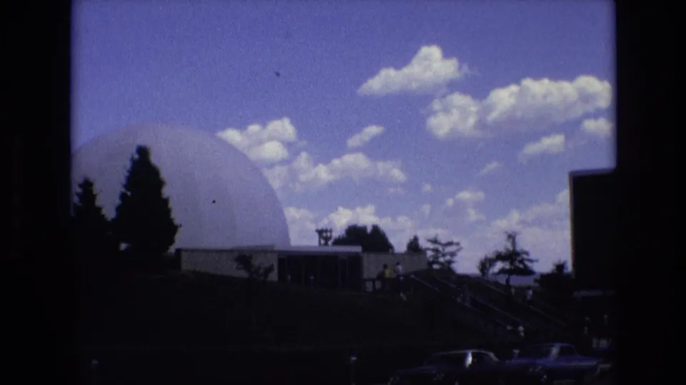 1973 Colorado Springs home-movie frame showing a large white geodesic dome near dark building sections, silhouetted evergreens, stairs, cars, and a bright clouded sky.