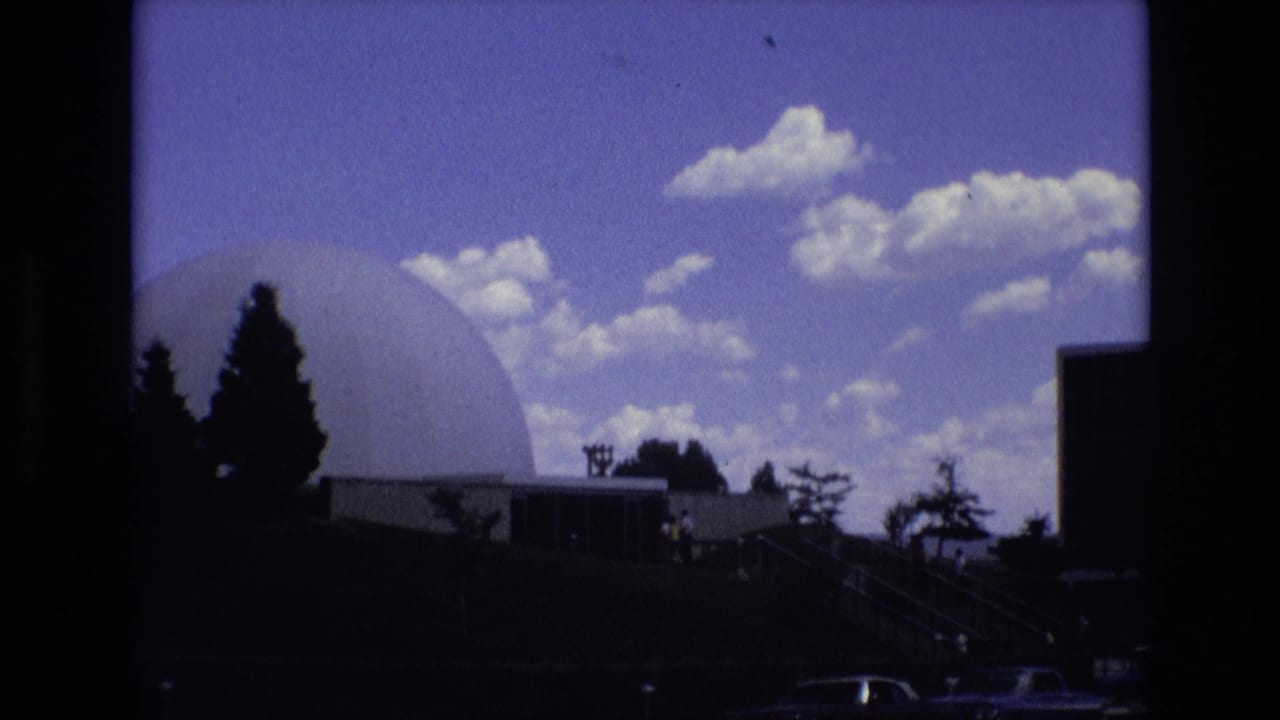 Dome structure beside trees and a building, with cars along the lower edge.