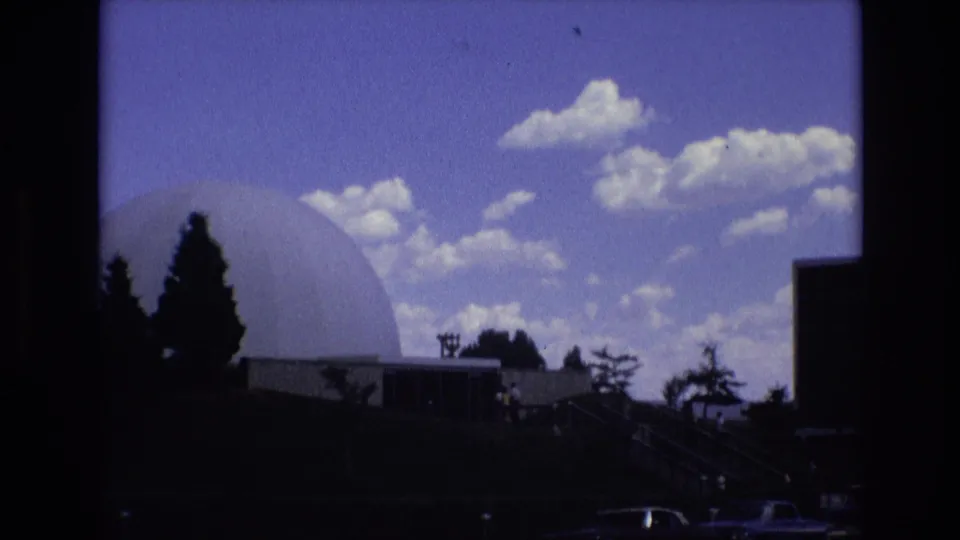 1973 Colorado Springs frame with the white dome partly framed by dark vertical edges, evergreen silhouettes, low building sections, parked cars, and bright clouds overhead.