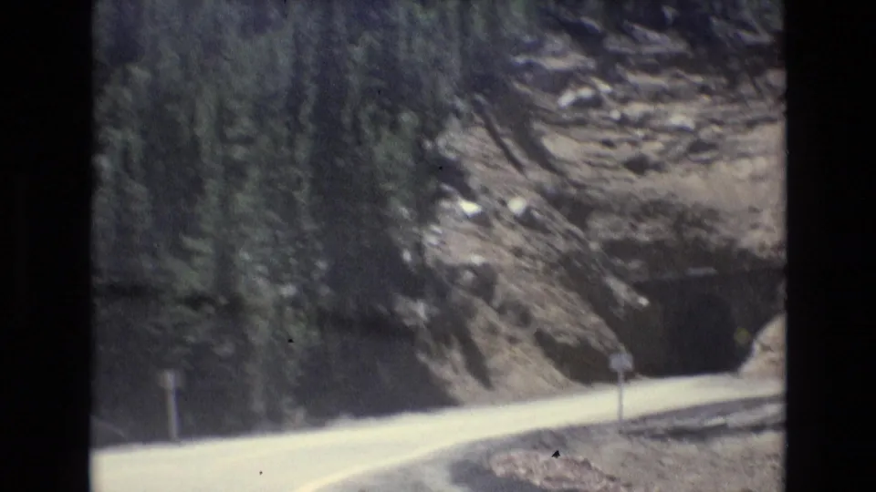 1973 Colorado Springs home-movie frame showing a pale road curving beside a rocky wooded slope, a dark roadside sign, and soft 8mm motion blur.