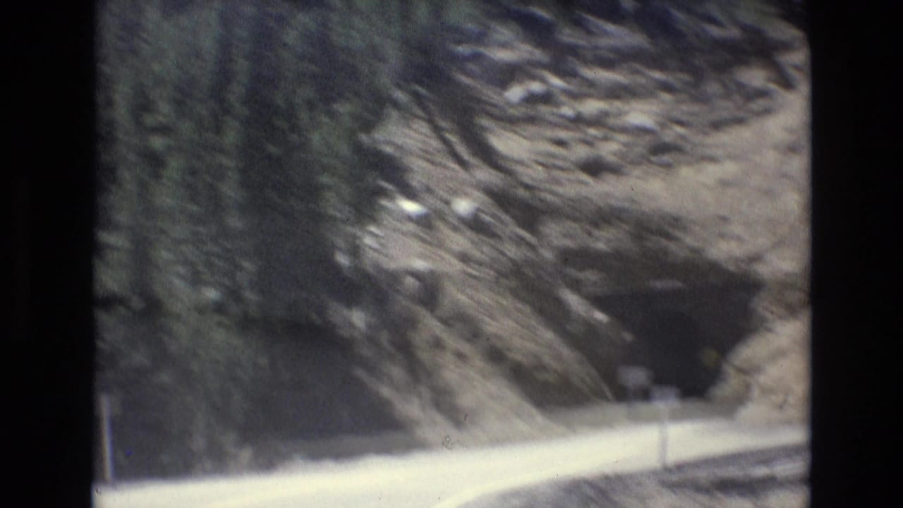 1973 8mm home-movie frame from Colorado Springs showing a paved mountain road approaching a dark rocky tunnel with forested slopes above.