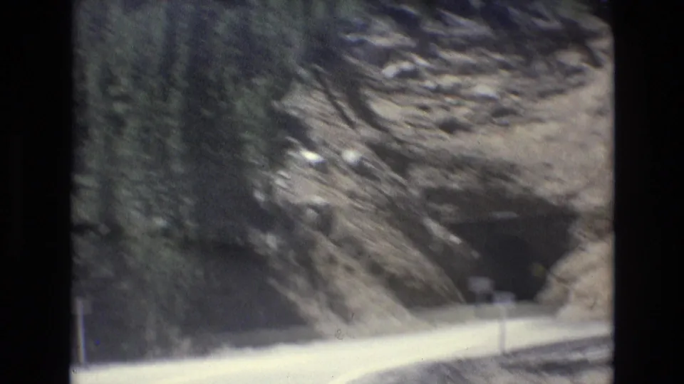 1973 8mm home-movie frame from Colorado Springs showing a paved mountain road approaching a dark rocky tunnel with forested slopes above.