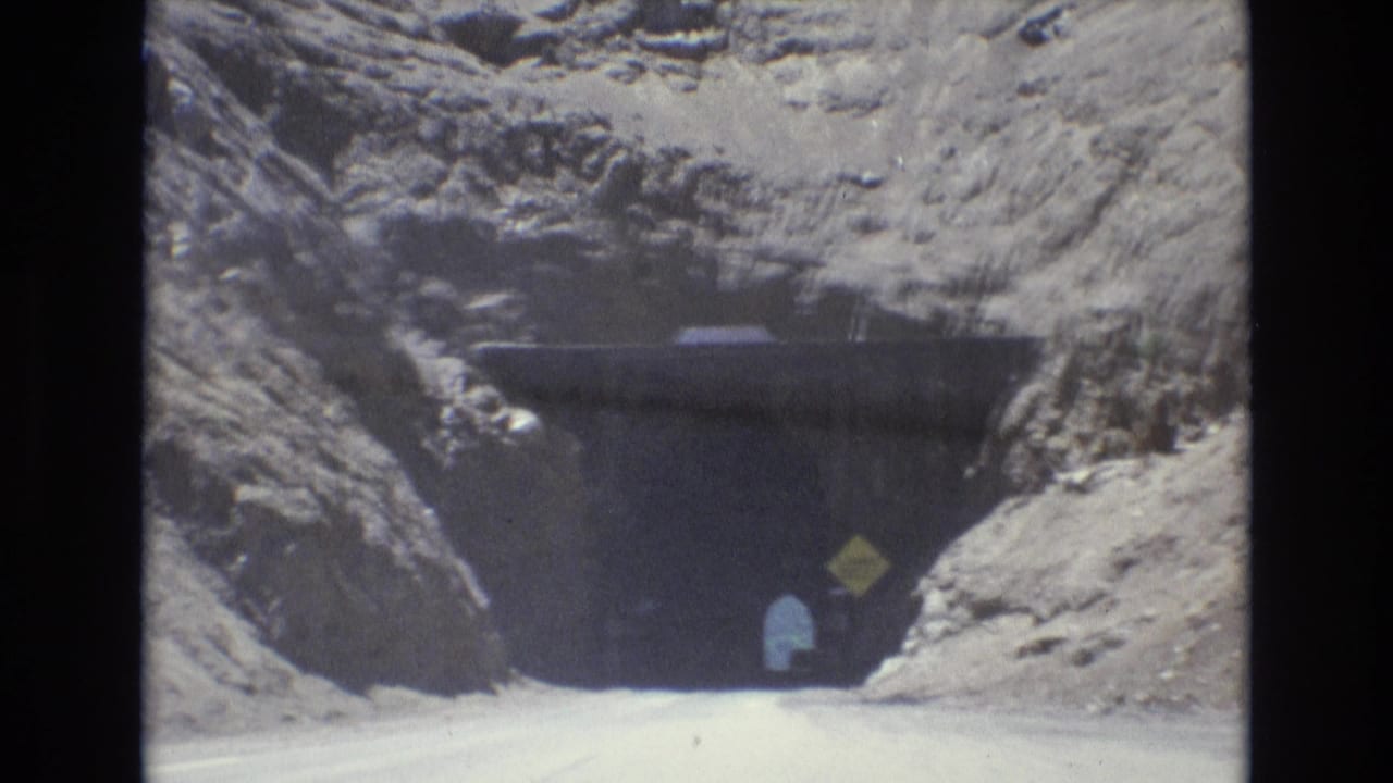 1973 8mm frame from Colorado Springs showing a road approaching a dark tunnel in pale rocky terrain, with a yellow clearance sign and a small figure near the entrance.