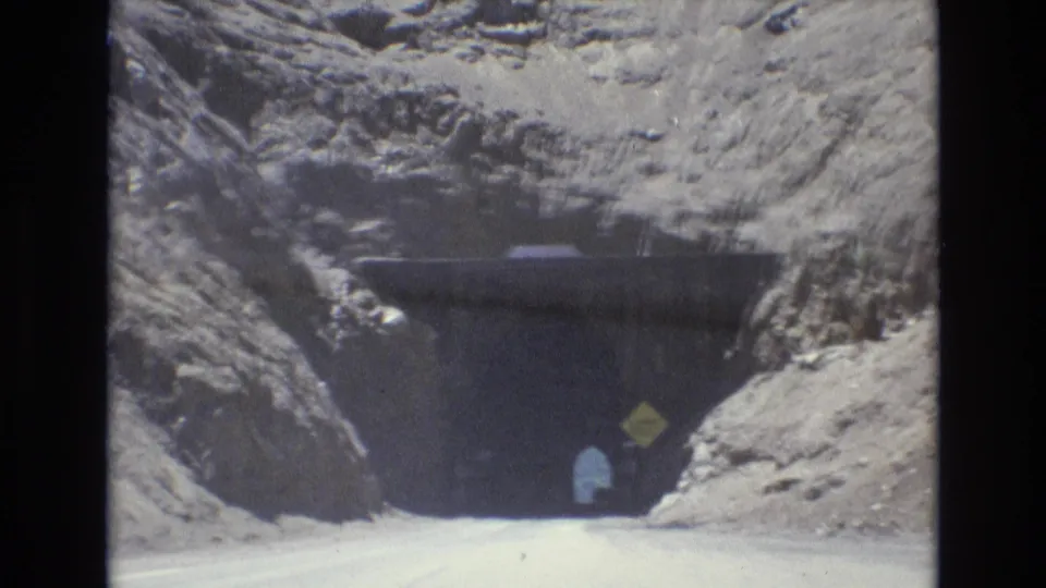 1973 8mm frame from Colorado Springs showing a road approaching a dark tunnel in pale rocky terrain, with a yellow clearance sign and a small figure near the entrance.