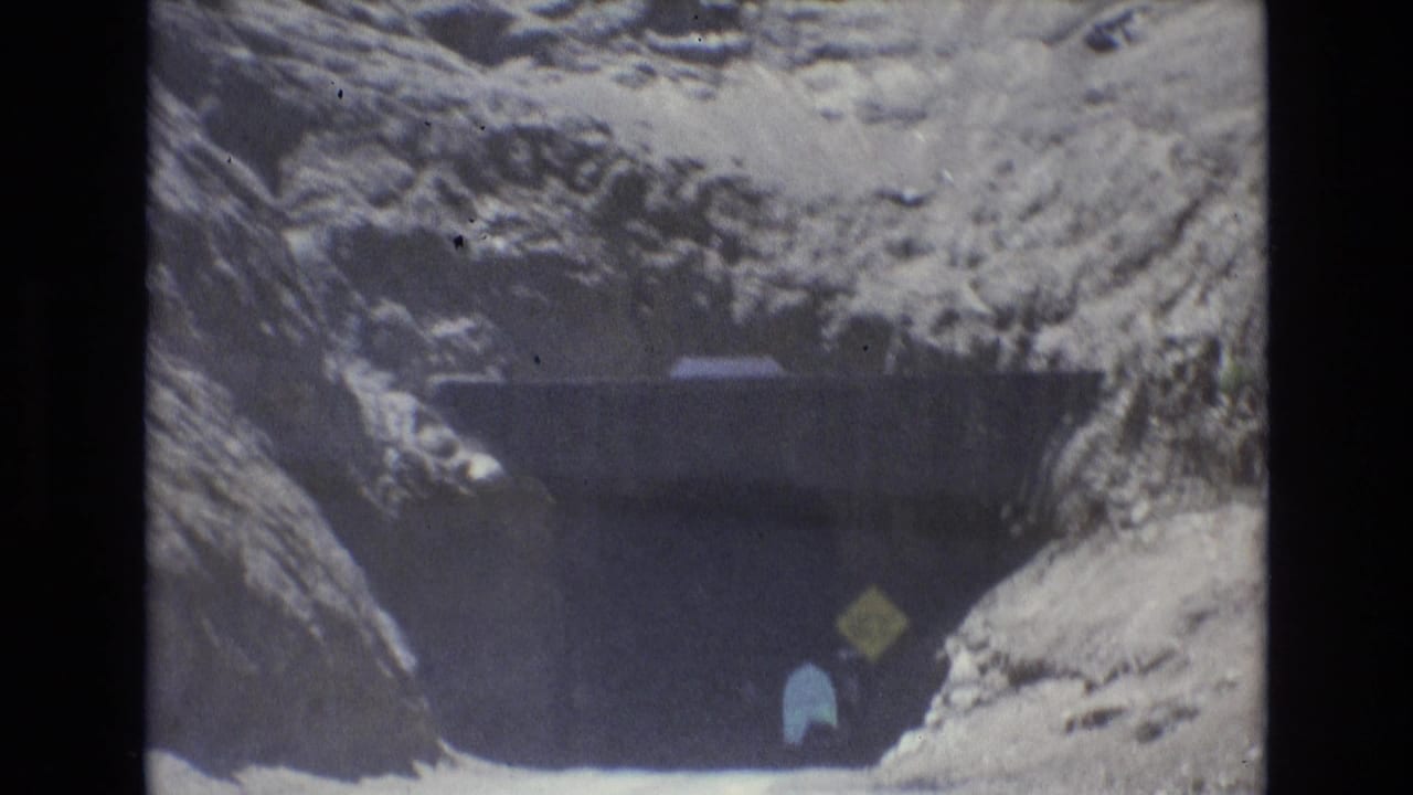 Dark tunnel entrance in rocky terrain with a yellow sign and small figure near the roadway.