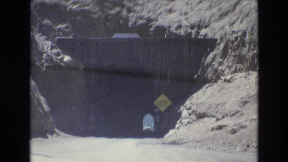 Closer 1973 8mm view of a Colorado Springs mountain tunnel entrance, showing rough rock, a dark opening, a yellow sign, and a small figure near the road edge.