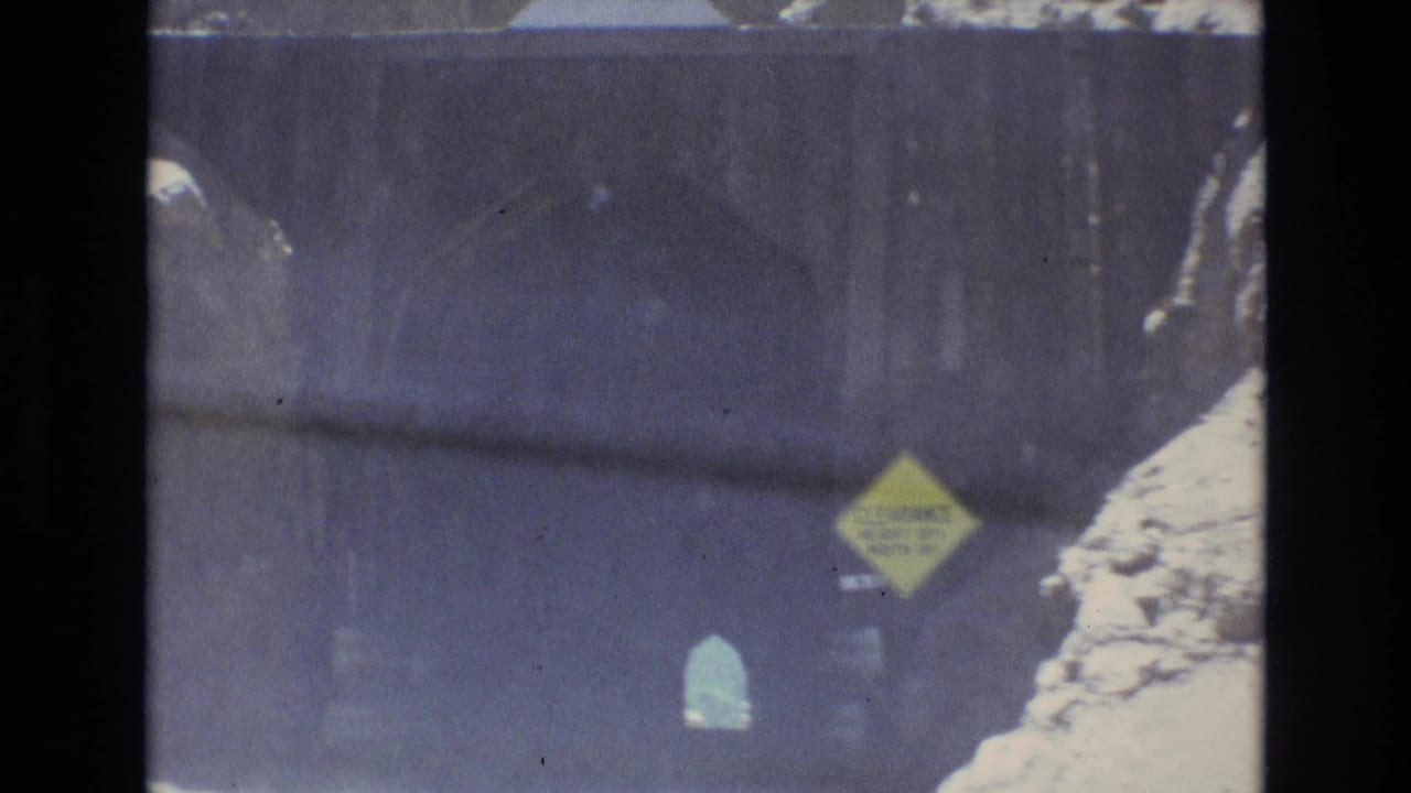 1973 Colorado Springs home-movie frame showing a car approaching a dark mountain tunnel, with a yellow clearance warning sign and rocky roadside visible through the windshield.