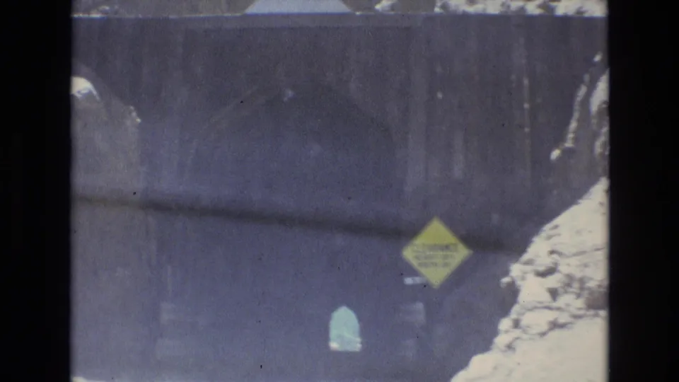 1973 Colorado Springs home-movie frame showing a car approaching a dark mountain tunnel, with a yellow clearance warning sign and rocky roadside visible through the windshield.