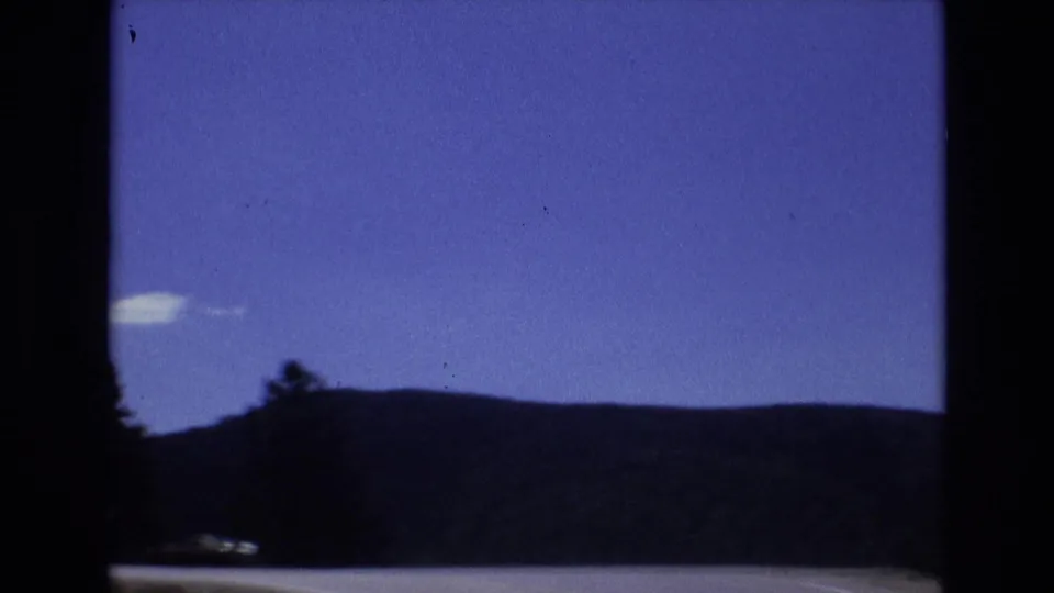 The car emerges from the tunnel into daylight, revealing blue sky, dark mountain silhouettes, the tunnel edge, and a faint yellow sign near the upper right.