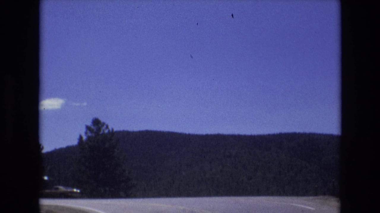 1973 Colorado Springs home-movie frame showing a moving roadside view with evergreen trees, dark wooded hills, and a deep blue sky.