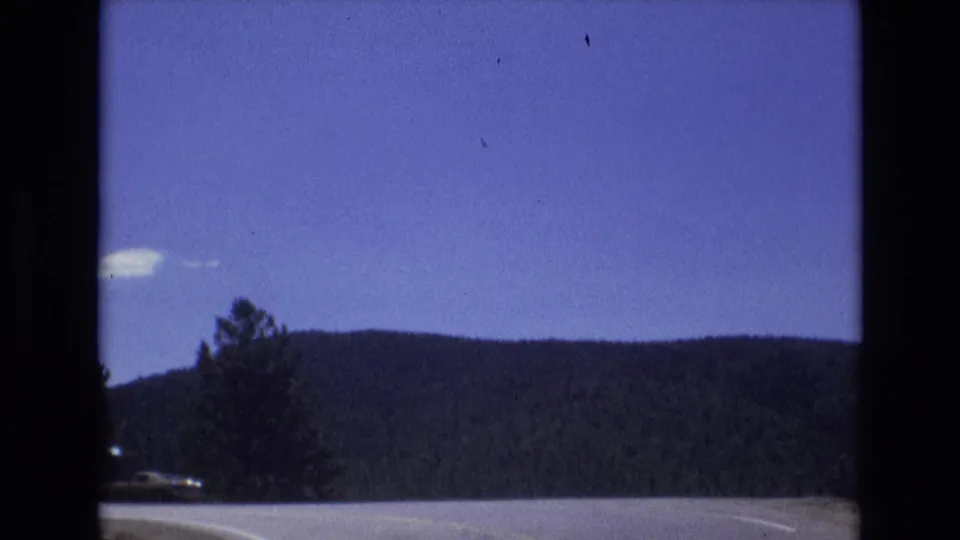 1973 Colorado Springs home-movie frame showing a moving roadside view with evergreen trees, dark wooded hills, and a deep blue sky.
