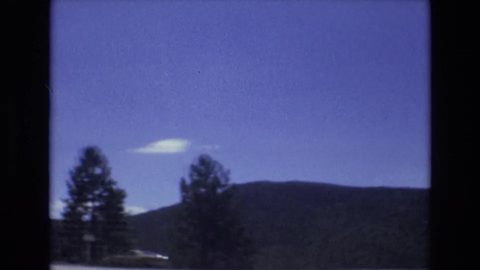 A 1973 Colorado Springs driving view frames two roadside evergreens, a rounded wooded hill, blue sky, and a small white cloud through dark 8mm borders.