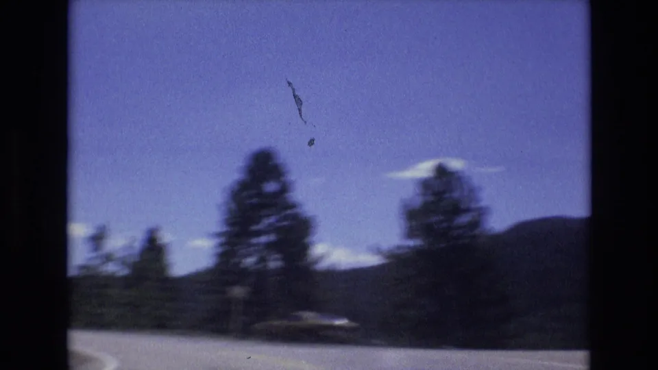 The moving 8mm camera catches blurred evergreen trees, a low dark hill, pale clouds, and roadside pavement during a 1973 Colorado Springs scenic drive.