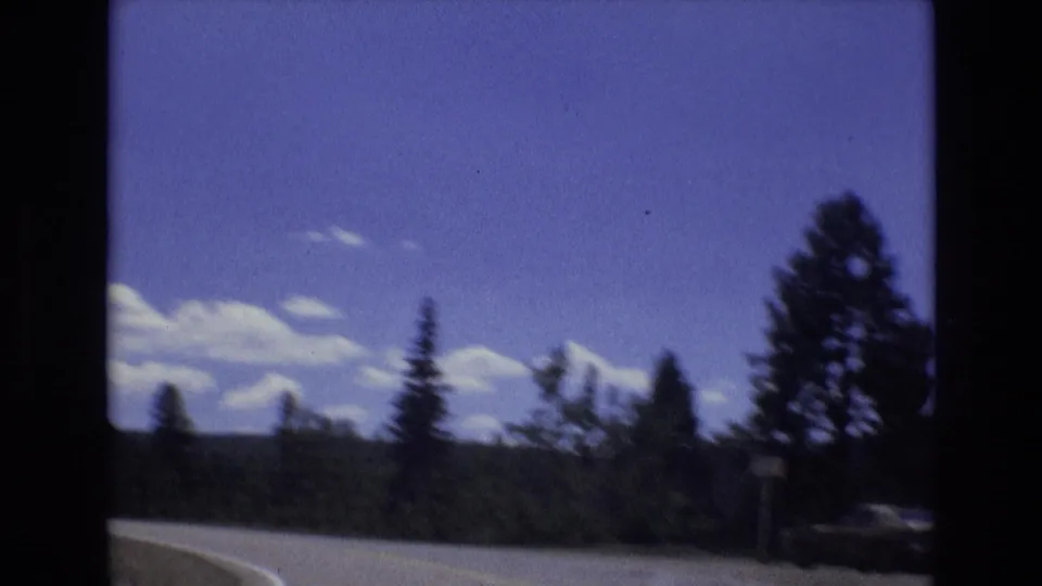 A 1973 Colorado Springs road view shows a curving paved road, evergreen silhouettes, layered white clouds, and distant wooded hills in saturated 8mm color.