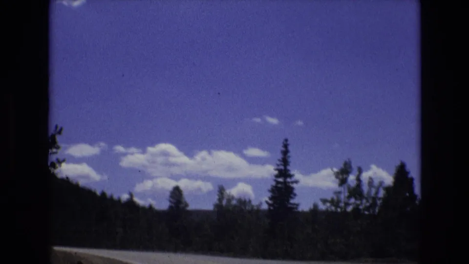 1973 8mm Colorado travel footage shows a winding road along forested hills beneath a blue sky with scattered white clouds.