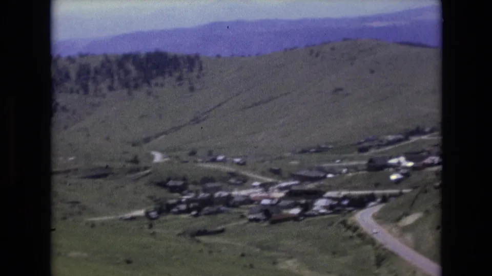 1973 8mm Colorado travel footage shows a small mountain town or settlement in a valley below rolling hills and distant mountains.
