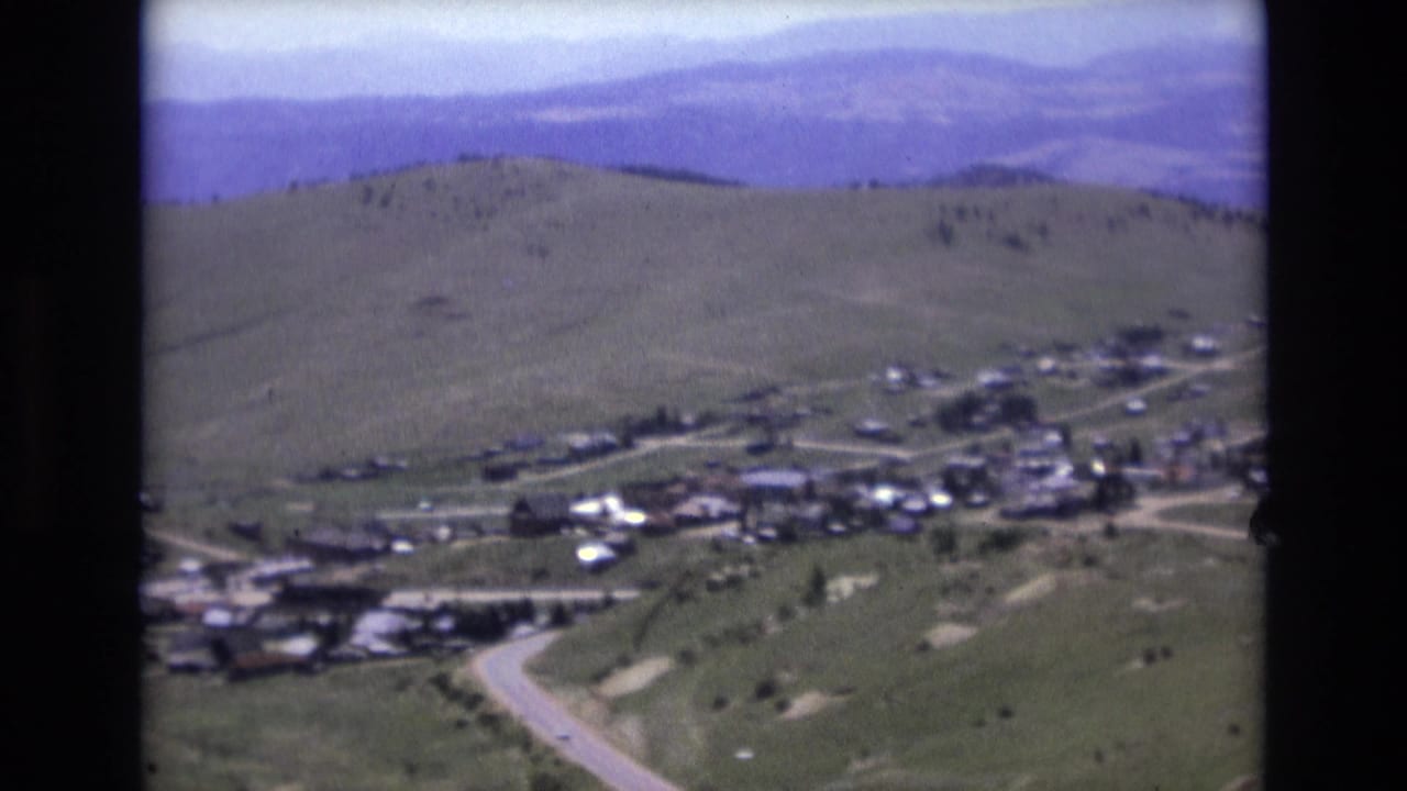 1973 8mm view near Colorado Springs showing a valley settlement, winding light roads, open green hills, and hazy blue-purple mountains in the distance.