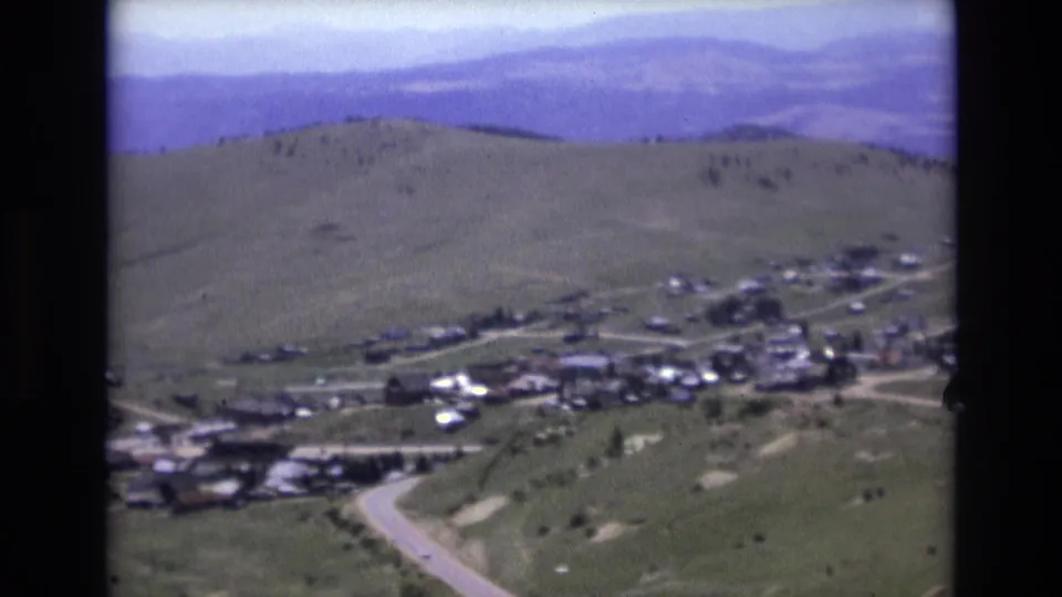 1973 8mm view near Colorado Springs showing a valley settlement, winding light roads, open green hills, and hazy blue-purple mountains in the distance.