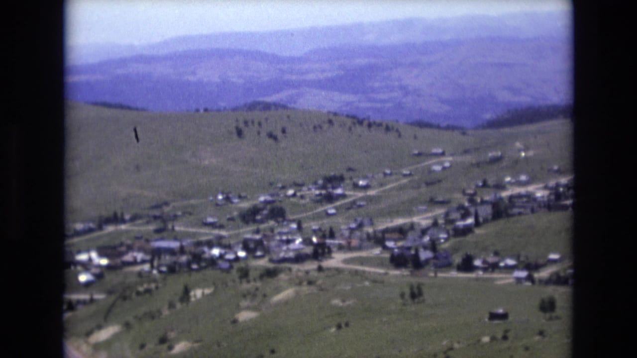 Village buildings and roads sit below rolling hills with layered mountains beyond.