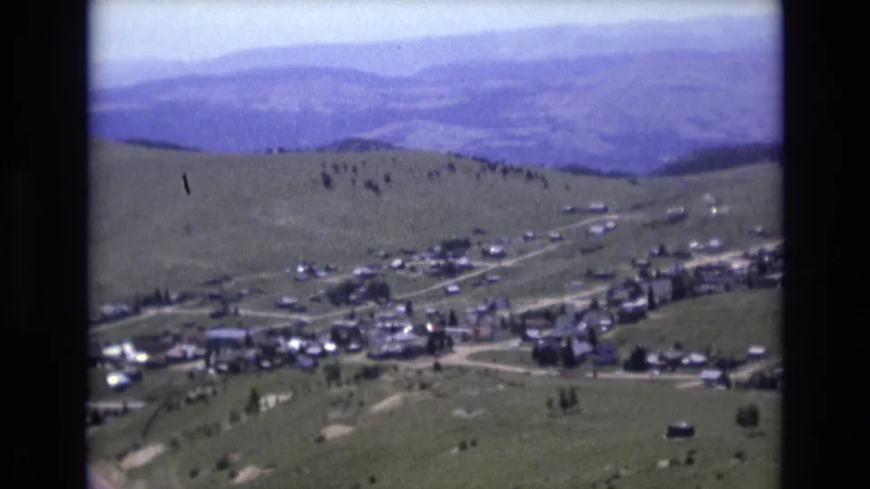 Elevated 1973 Colorado Springs-area home-movie frame with a small town cluster, light roads, sloping green hills, and layered mountain ridges beyond.