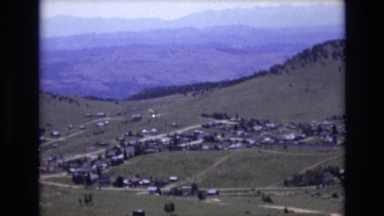 1973 8mm view near Colorado Springs showing a small settlement spread below rolling green foothills with hazy mountain ridges in the distance.