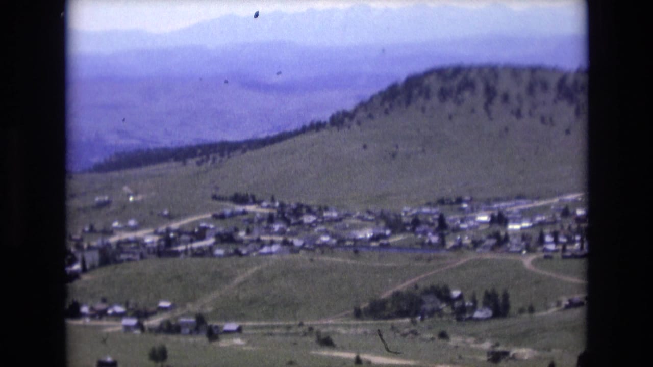 Elevated 1973 home-movie frame near Colorado Springs with clustered buildings, open green slopes, tree lines, and distant hazy mountains.