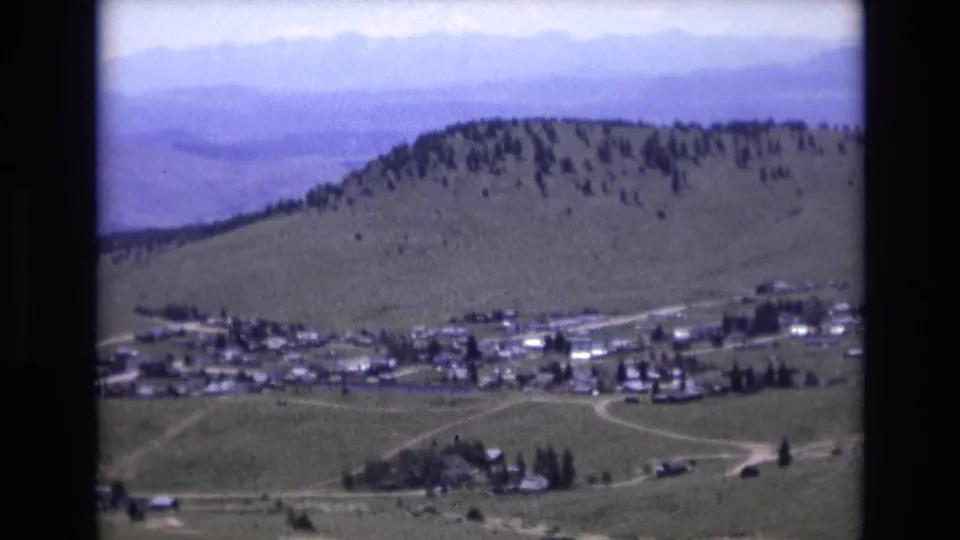 1973 Colorado Springs-area panorama showing a village across the lower frame, a rounded green hill with scattered trees, pale roads, and hazy mountain ranges beyond.