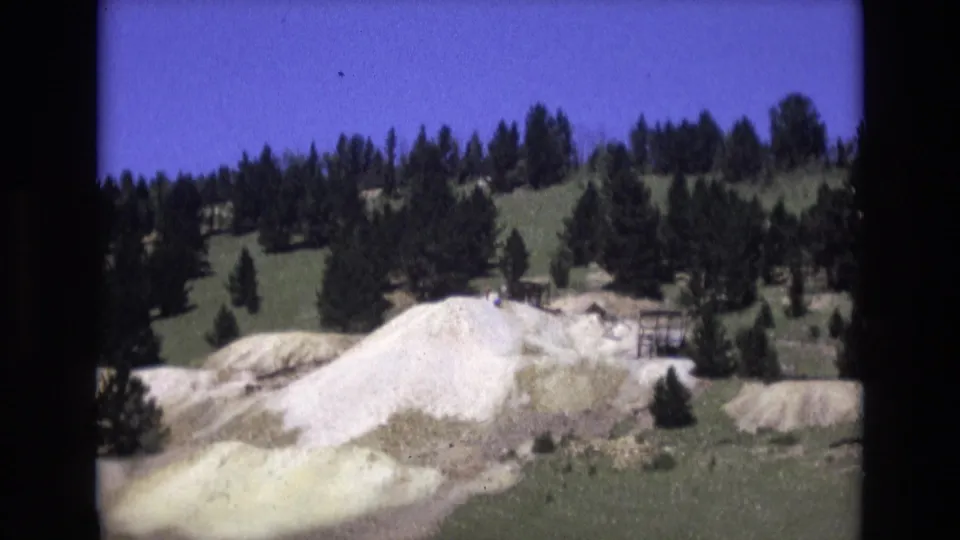 1973 Colorado Springs 8mm frame showing a pale white and yellowish rock face below a pine-covered hillside under a saturated blue sky.