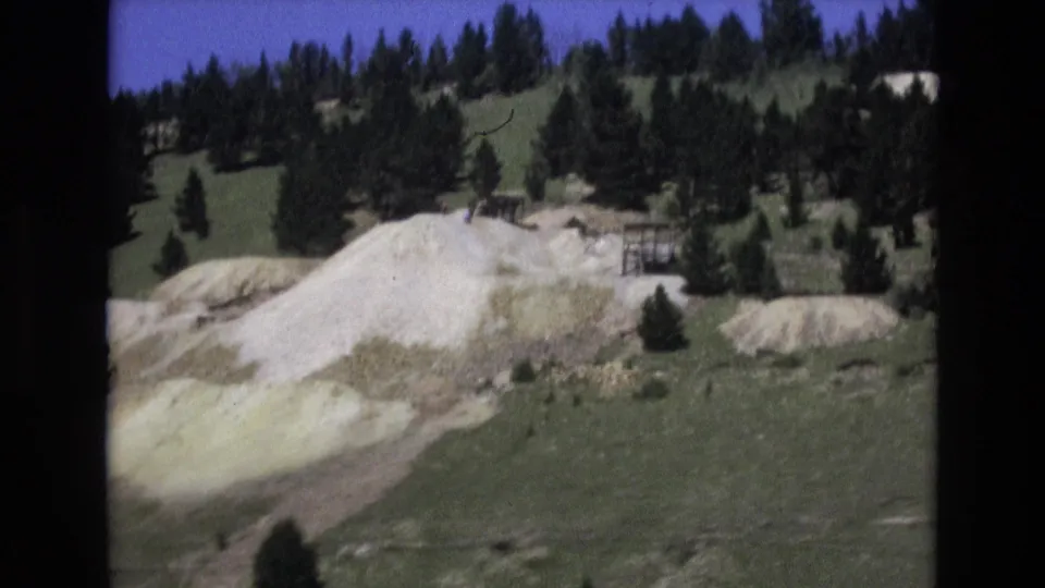 1973 home-movie frame of a white rock face in a grassy Colorado Springs hillside landscape, with dark pines along the upper slope.