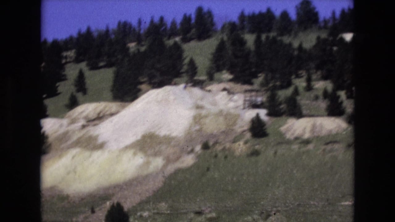 1973 Colorado Springs 8mm frame showing light-colored earth mounds below a tree-covered slope under a blue sky.