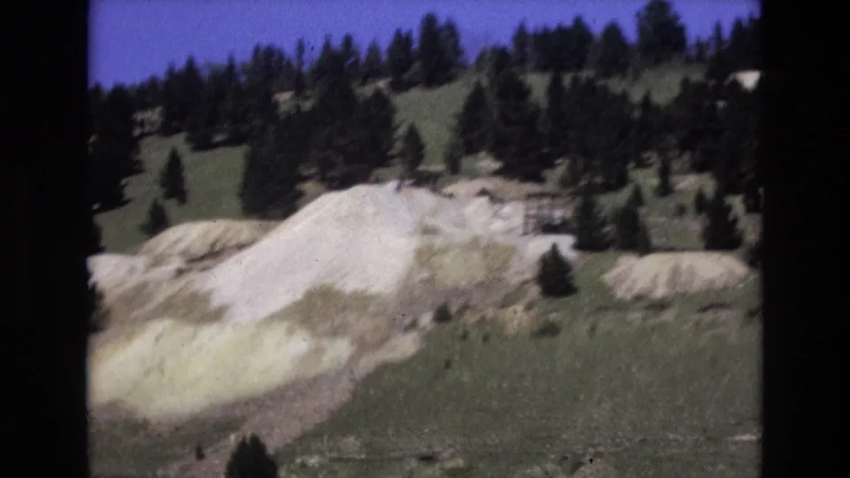1973 Colorado Springs 8mm frame showing light-colored earth mounds below a tree-covered slope under a blue sky.
