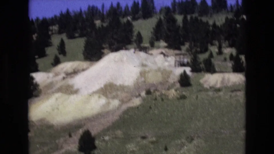 1973 Colorado Springs 8mm view of a pale mound across open ground, with scattered evergreens and a wooded slope in the background.