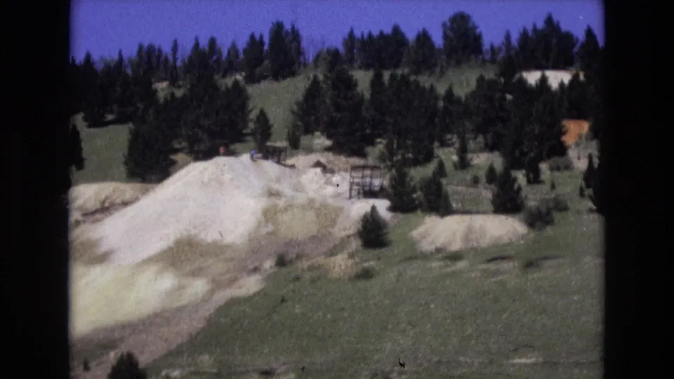 1973 Colorado Springs home-movie frame with a pale hillside deposit, green foreground, evergreen trees, and a small distant structure or fence.
