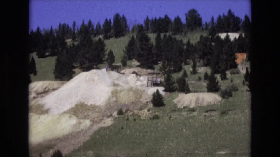 1973 Colorado Springs 8mm frame showing a pale sloped mound in open terrain, backed by evergreens and a wooded hillside.