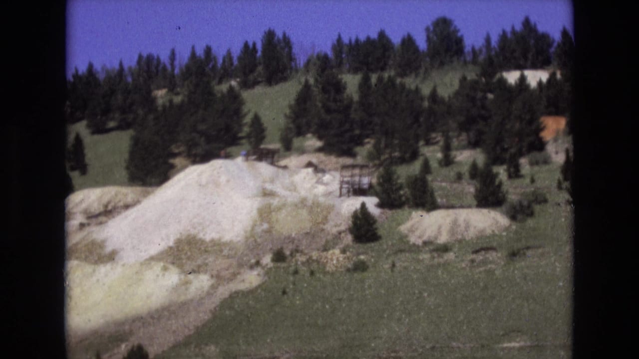 1973 Colorado Springs-area home-movie frame showing a green hillside with conifer trees, pale exposed rock or gravel, and a small dark structure or sign near the slope.