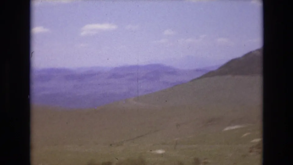 Wide 1973 Colorado home-movie view of an open hillside or valley edge with layered distant mountain ranges and a hazy blue sky.