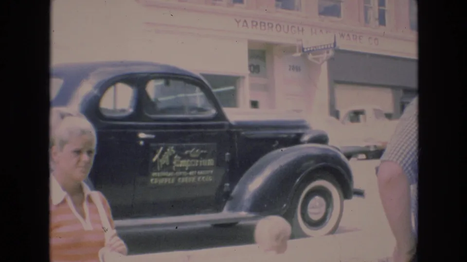 1973 Colorado Springs downtown frame showing a black coupe with door lettering and whitewall tires outside Yarbrough Hardware Co., with a woman, child, and another person partly visible.