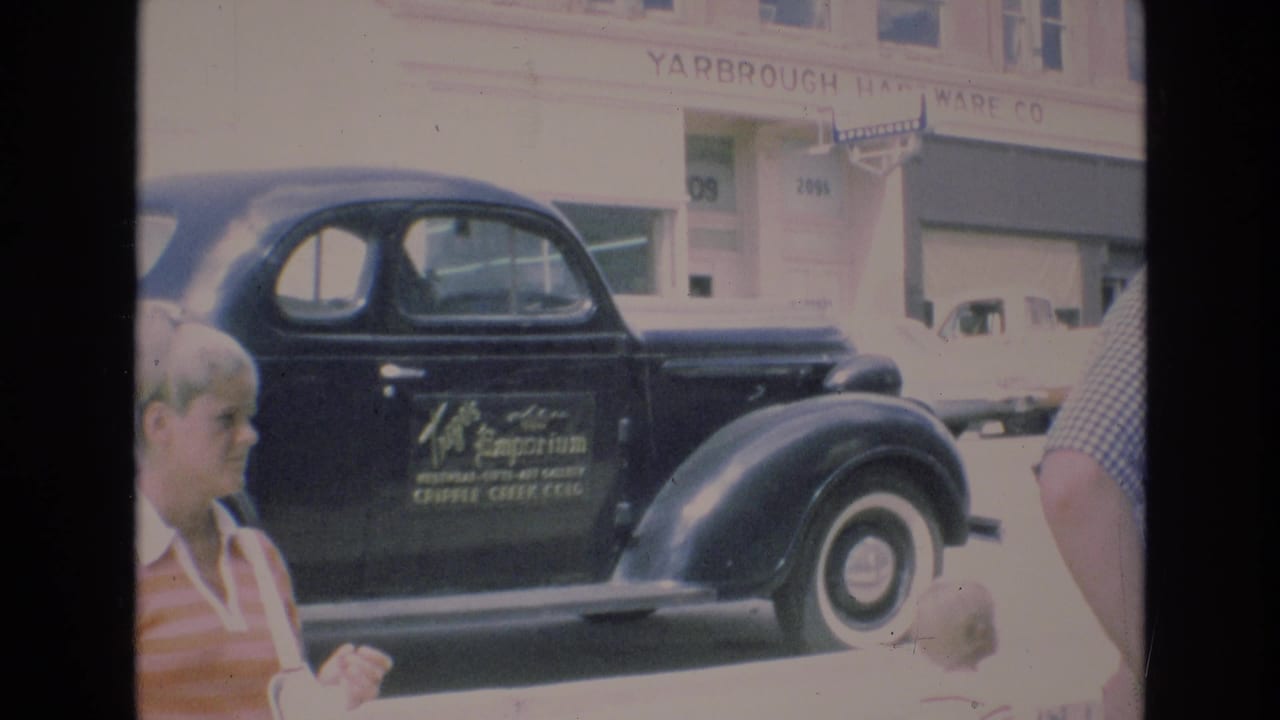 1973 Colorado Springs street view showing a black vintage sedan with Trego's Emporium lettering, pedestrians in the foreground, and Yarbrough Hardware Co. signage in the background.