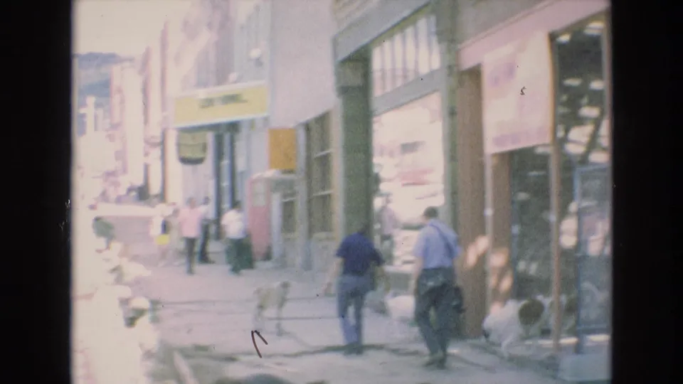 1973 Colorado Springs sidewalk scene with pedestrians moving past storefront windows and signs in a softly exposed 8mm home-movie frame.